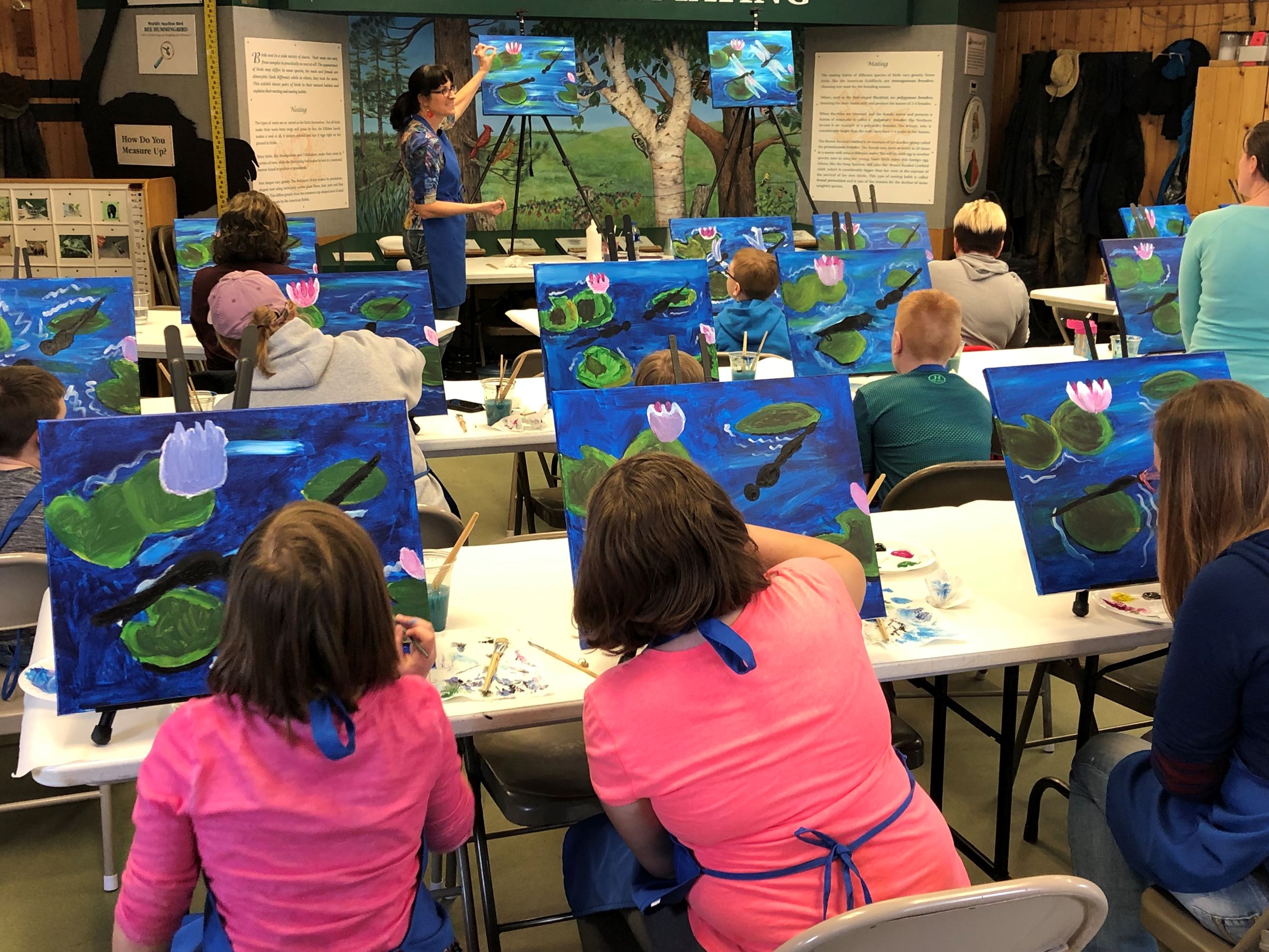 Ten people doing a guided painting class. The instructor is at the front of the room showing people 
