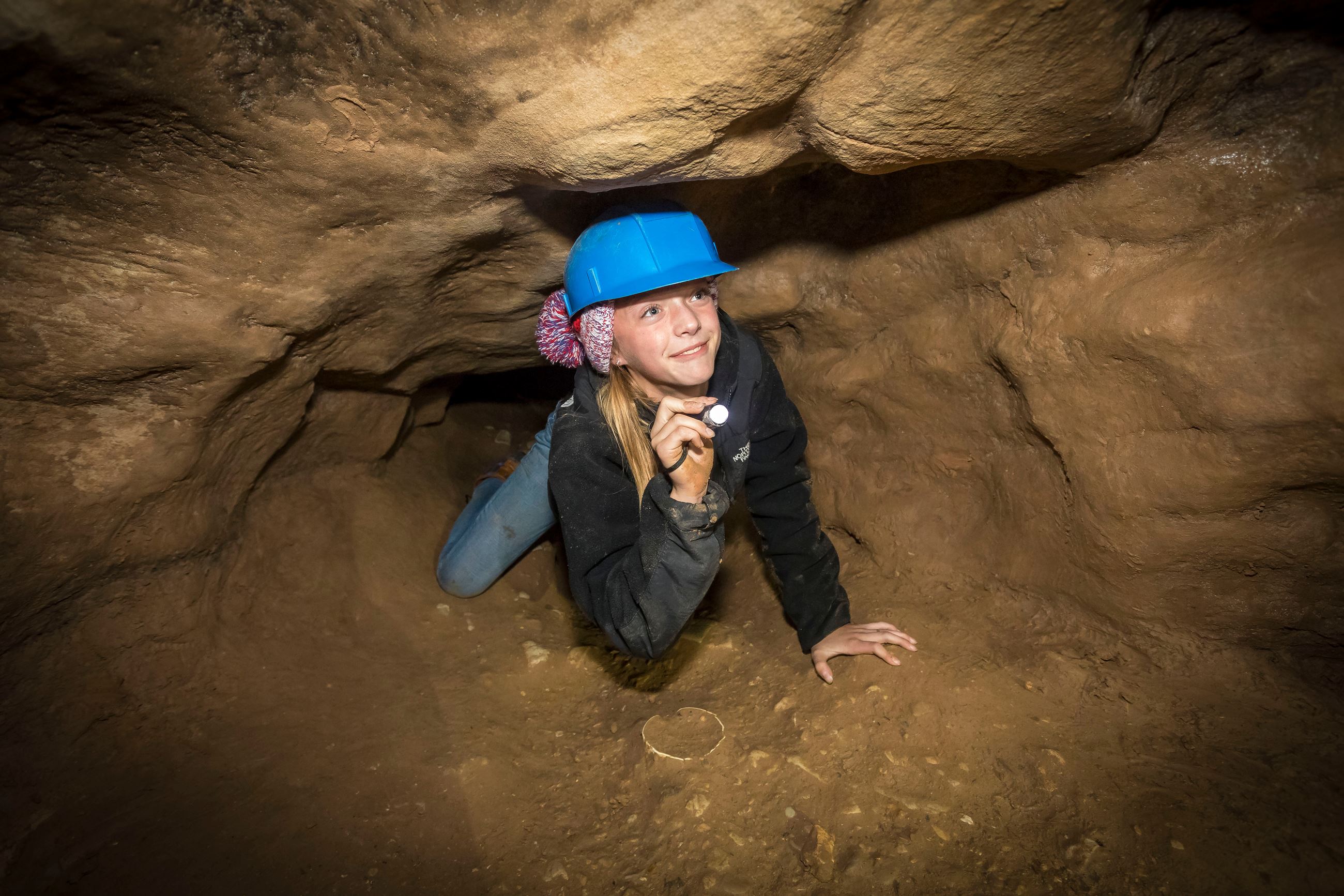 A girl wearing a blue helmet and holding a flashlight is crawling on her hands and knees through a t