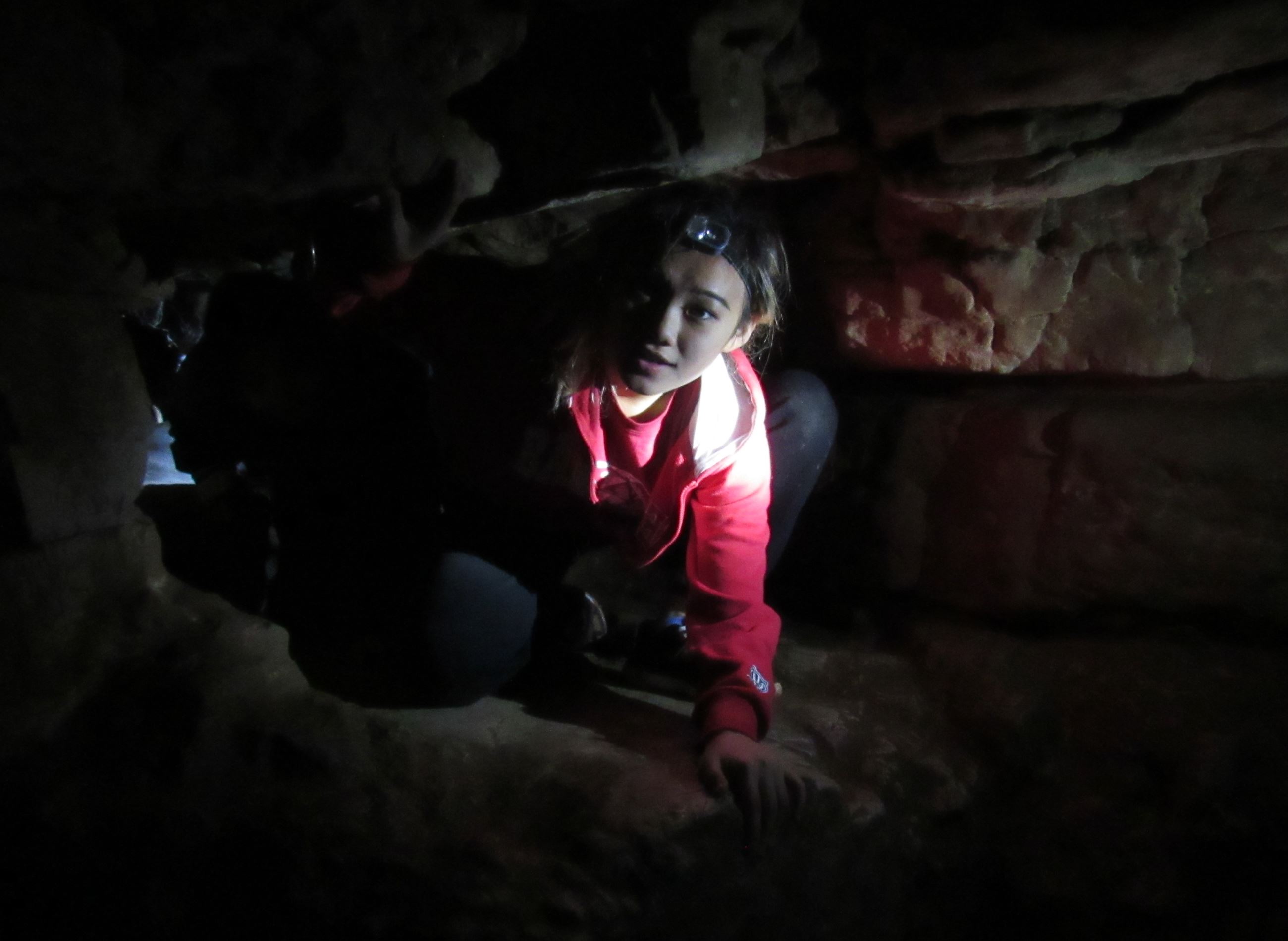 A teenage girl wearing a red sweatshirt and a headlamp crawling through a tunnel in a cave.